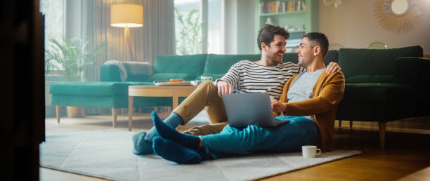 istock Handsome Gay Couple Using Laptop Computer, while Sitting on a Living Room Floor in Cozy Stylish Apartment. Adult Boyfriends Online Shopping on Internet, Watching Funny Videos on Streaming Service. 1344243615