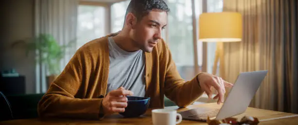 Photo of Handsome Adult Man Using Laptop Computer, Sitting in Kitchen, Eating Breakfast Porridge or Cereal in Apartment. Attractive Man is Online Shopping, Watching Funny Videos on Streaming Service.
