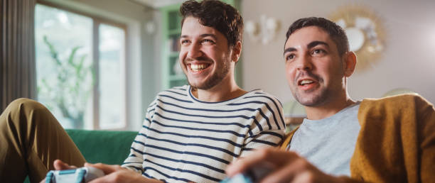 istock Happy Gay Couple Sitting on the Sofa Playing Video Games, Using Controllers. Competititve LGBT Men in Love have Fun Playing Online Video Games Together at Home in Stylish Apartment. 1344243621
