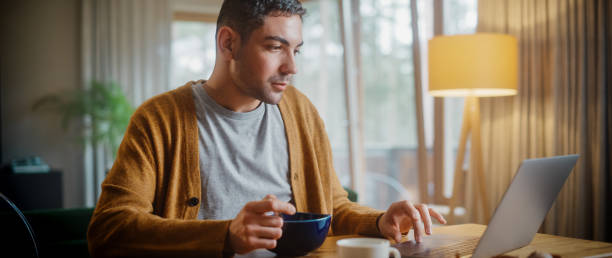 istock Handsome Adult Man Using Laptop Computer, Sitting in Kitchen, Eating Breakfast Porridge or Cereal in Apartment. Attractive Man is Online Shopping, Watching Funny Videos on Streaming Service. 1344243638