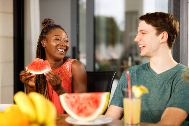 Happy couple talking while eating watermelon on a patio. Happy African American woman talking to her Caucasian boyfriend while eating watermelon on a patio. black man eating watermelon stock pictures, royalty-free photos & images