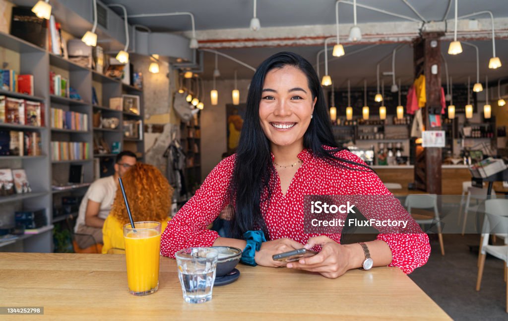 Cheerful and modern female entrepreneur of Asian ethnicity at the coffee shop, surfing the net on mobile phone Portrait of a modern female entrepreneur, at the coffee shop, surfing the net on a mobile phone and drinking coffee Mongolian Ethnicity Stock Photo