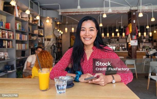 Cheerful And Modern Female Entrepreneur Of Asian Ethnicity At The Coffee Shop Surfing The Net On Mobile Phone Stock Photo - Download Image Now
