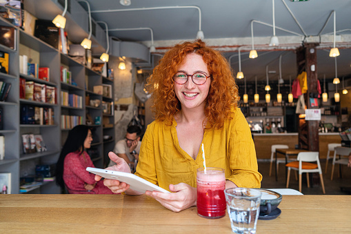 Portrait of a charming and modern young redhead woman, e-learning via digital tablet, at the modern and cozy cafeteria