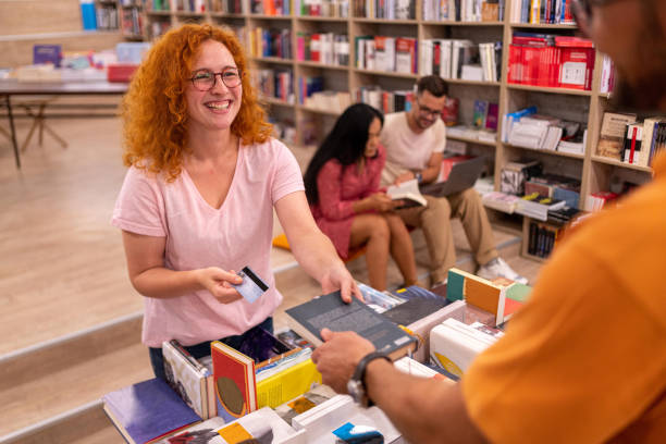 aufgeregte rothaarige junge frau, die mit einer kreditkarte für den buchkauf in der modernen buchhandlung bezahlt - bookstore stock-fotos und bilder