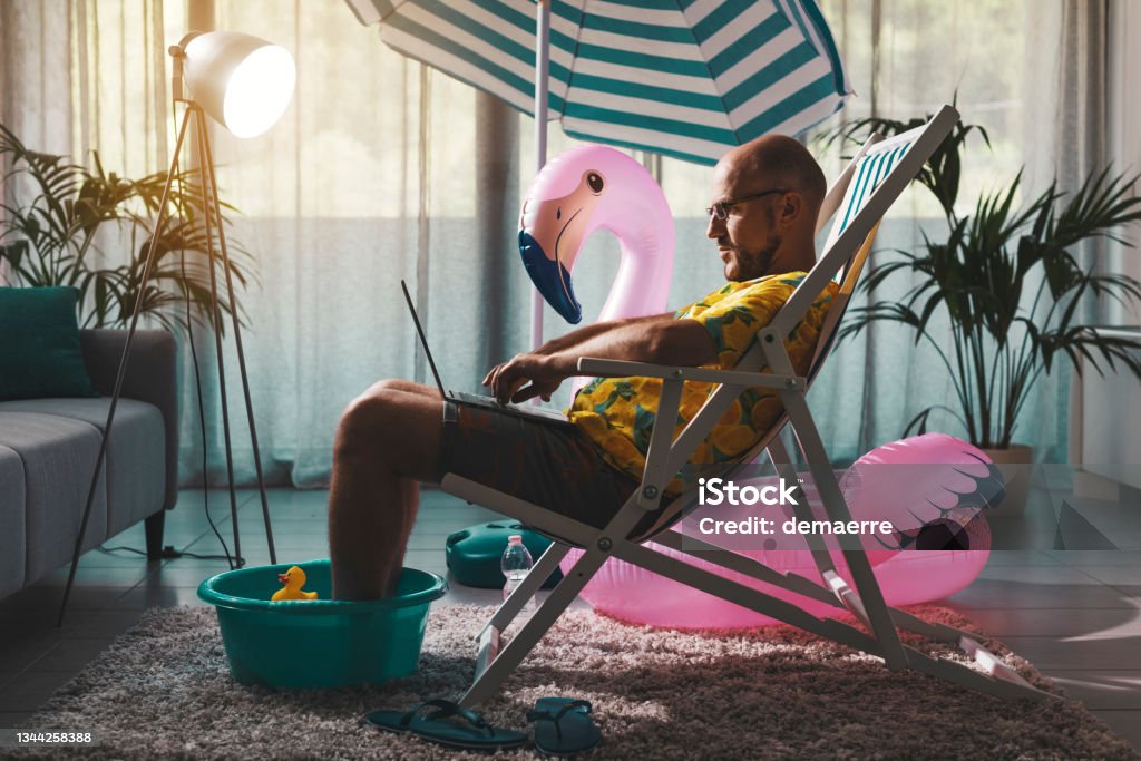 Man working from home during summer Man spending summer vacations at home alone, he is sitting on the deckchair in the living room and working with a laptop Working At Home Stock Photo