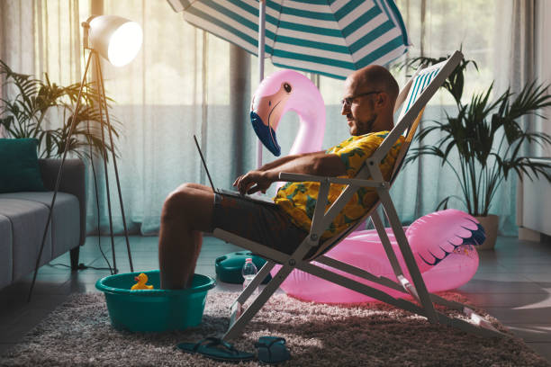 Man working from home during summer Man spending summer vacations at home alone, he is sitting on the deckchair in the living room and working with a laptop stay at home order stock pictures, royalty-free photos & images