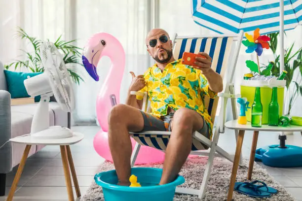 Happy man spending summer vacations at home and pretending he is on a beach, he is sitting on a deckchair and taking selfies with his smartphone