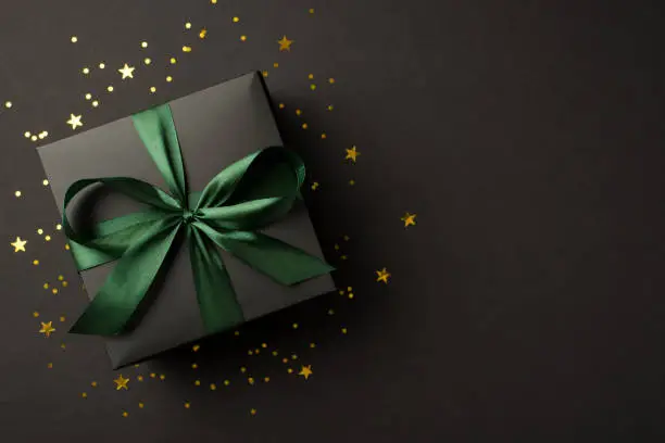 Photo of Top view photo of stylish giftbox with green ribbon bow golden stars and confetti on isolated black background with copyspace