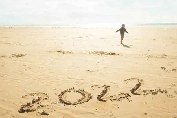 Year 2022 written in the sand of a beach. A child is running happily at the background. Peaceful and positive image.