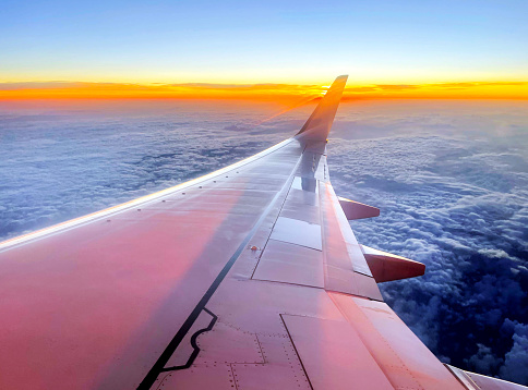 In flight view along the aluminum skinned wing of a modern jet airliner as the sun rises at dawn, showing aerodynamic control surface details of flaps, ailerons, airbrakes and winglets, looking towards the horizon with colourful sunrise effects and dense cumulus cloud cover below