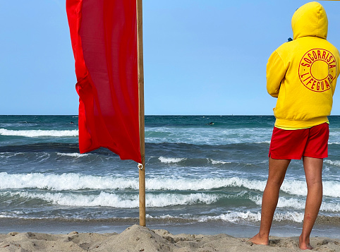 Lifeguard on Son Serra de Marina beach, Majorca, Balearic Islands, Spain. Spanish Lifeguard or Socorrista stands guard next to red flag warning on windswept Majorcan beach where dangerous currents and tides are a hazard to swimming, Balearic Islands, Spain