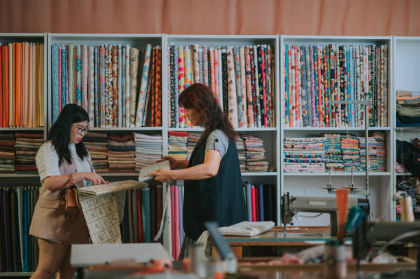 donna anziana cinese asiatica che raccoglie e mostra al suo cliente la scelta dell'abbigliamento in tessuto su un nuovo suggerimento di abbigliamento su misura nel suo negozio - negozio di tessuti foto e immagini stock