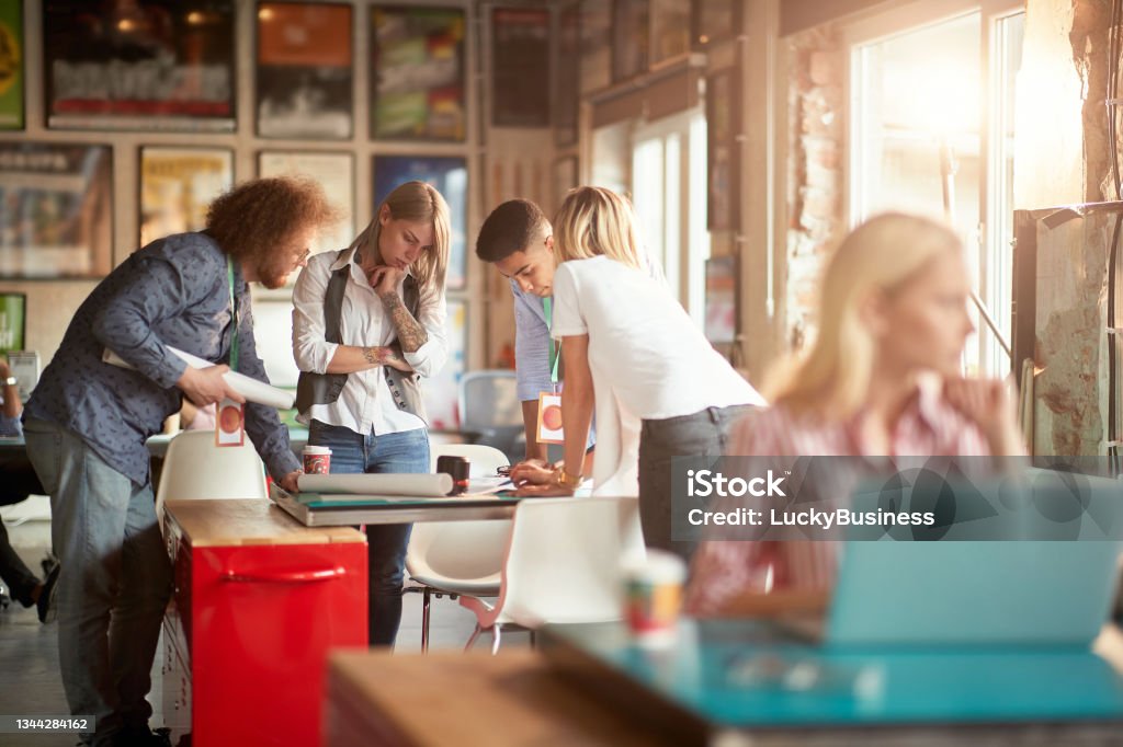 selective focus on group of young adult people gathered around the table, analyzing new design with teamleader selective focus on group of young adult people gathered around the table, analyzing new design Real Estate Office Stock Photo