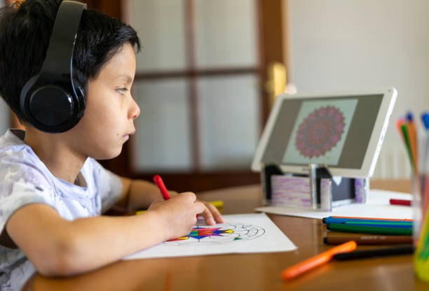 Child painting a mandala with his colored markers watching a tutorial on his tablet with headphones at home stock photo