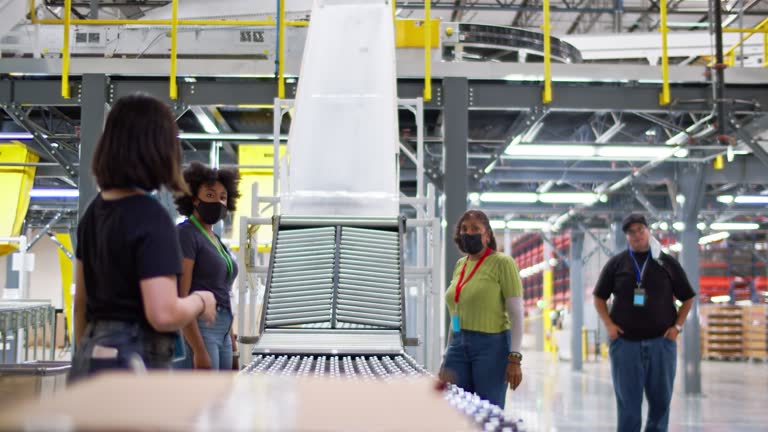 A diverse group of masked men and women working in a massive fulfillment center guide packages along a flexible conveyor belt at the bottom of a chute and into a truck pulled up at a loading dock.