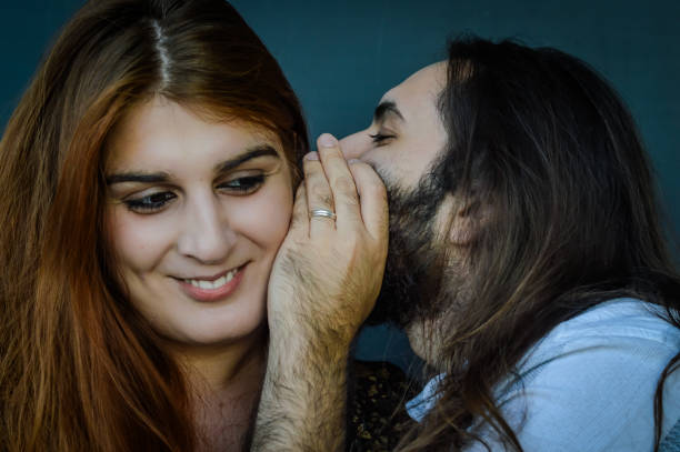 portrait of a young couple where bearded man with long hair tells a funny secret to the woman with red hair and she smiles - long hair red hair women men imagens e fotografias de stock