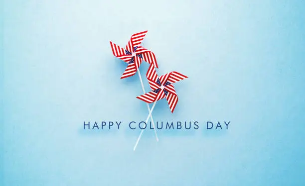 Photo of Happy Columbus Day Message and Paper Pinwheel Pair Textured with American Flag on Blue Background