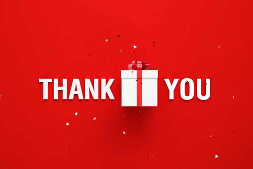 Thank you message and white gift box sitting behind gold colored confetti over red background. Horizontal composition with copy space. Directly above. Great use for Christmas concepts.
