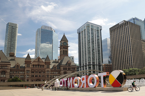Toronto, Canada - August 30, 2021: A cyclist walks their bicycle past the 3D TORONTO sign at Nathan Phillips Square. A bright medicine wheel, installed in 2018,  marks National Indigenous Peoples Day. Artist Danilo Deluxo McCallum designed the TORONTO wrap, Patterns of the People, in honour of International Decade for People of African Descent. \n\nBackground shows Old City Hall and clock tower in the Romanesque style . Modern towers line Bay Street in the Financial District.