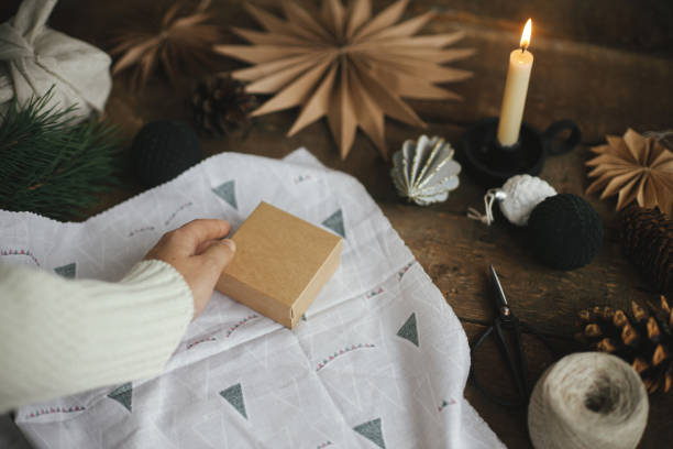 istock Hands wrapping christmas gift box in modern fabric on rustic wooden table with scissors, craft paper star, candle. Zero waste and eco friendly presents. Atmospheric moody time, nordic style. 1344304152