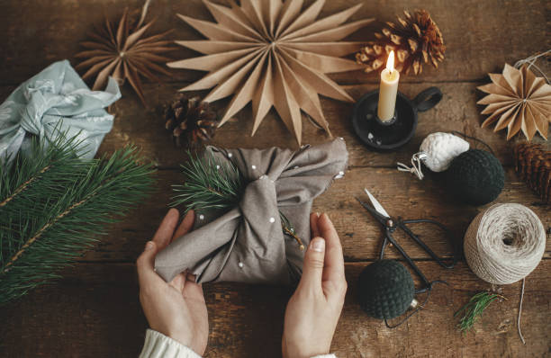 istock Christmas gift rustic flat lay. Hands holding stylish xmas gift wrapped in brown fabric on rustic wooden table with scissors, paper star, candle. Atmospheric moody image. Furoshiki wrap, zero waste 1344304736