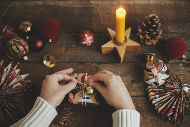 istock Hands decorating wrapped gift box with golden ornament on rustic wooden table with candle, scissors, red paper star. Atmospheric moody image. Merry Christmas! Winter Holidays preparation 1344304855