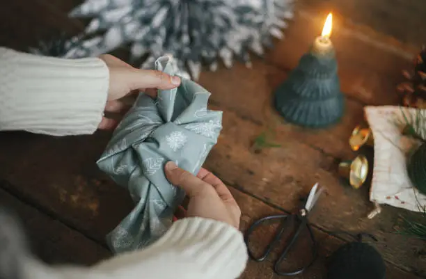 Photo of Hands wrapping christmas gift in fabric on rustic wooden table with candle, scissors, blue paper stars. Atmospheric moody image, nordic style. Merry Christmas and Happy Holidays! Furoshiki wrap