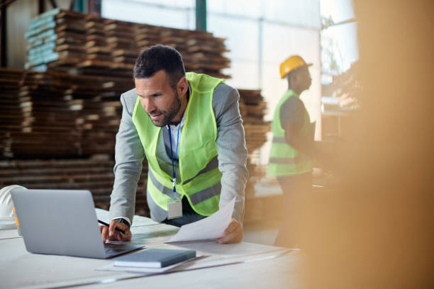 Young businessman using laptop while making distribution plans at lumber warehouse. Warehouse manager working on laptop while going through paperwork at lumber distribution department. Copy space. foreman stock pictures, royalty-free photos & images