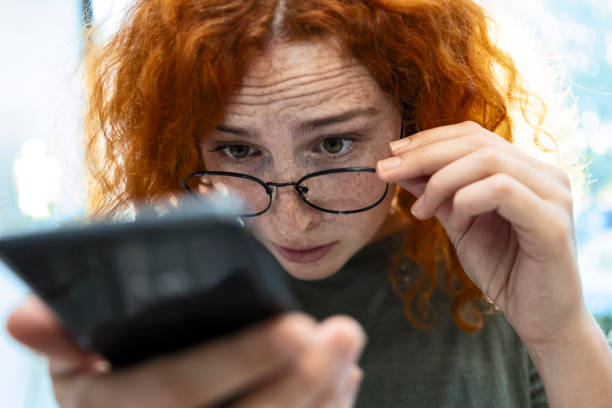 Redhead young woman with eyesight problems trying to read smart phone text