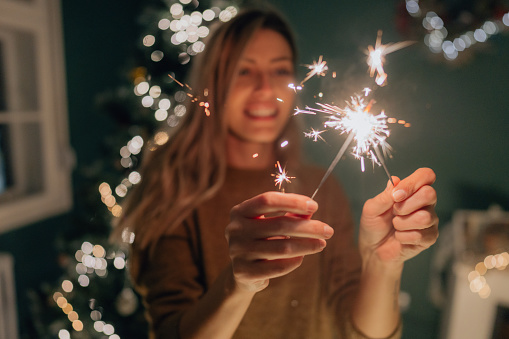 Photo of a smiling woman holding sparklers and enjoying the lovely Christmas eve in the living room of her apartment;