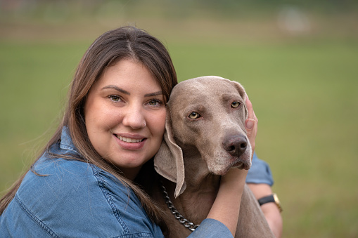 face of a beautiful young woman and a weimaraner dog together looking at the camera outdoors