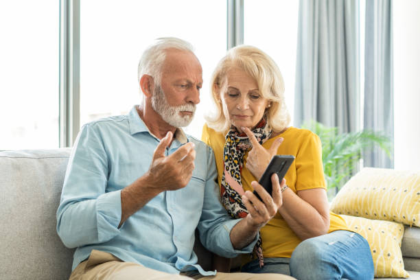 Worried senior couple reading bad news on phone. Worried senior couple using phone while sitting on sofa at home. couple isolated wife husband stock pictures, royalty-free photos & images