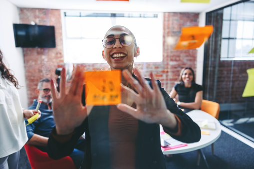 Smiling businesswoman sticking adhesive notes to a glass wall with her colleagues in the background. Happy young businesswoman sharing her ideas with her team in a modern office.