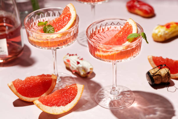 champagne, rose sparkling wine or paloma cocktails in crystal glasses on pink background with mini eclairs. refreshing beverage with grapefruit slice and mint. summer drinks, selective focus - pink champagne fotos imagens e fotografias de stock