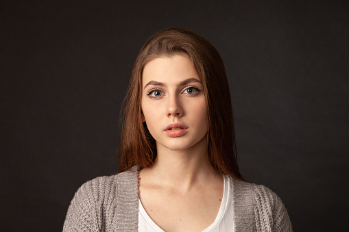 Close up studio portrait of attractive 19 year old woman with long brown hair in gray cardigan sweater on black background