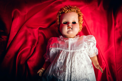 A creepy old baby doll with her eyes closed laying on a red satin sheet.