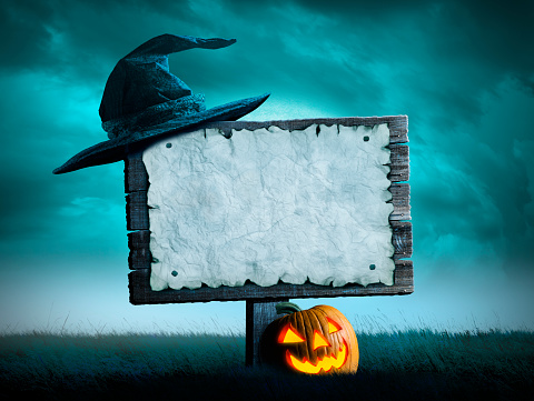 A witch's hat hangs from the corner of an old wooden sign that has a blank sheet of paper posted to it.  An illuminated jack o'lantern rests at the base of the sign that stands in a grassy field on a spooky Halloween night.