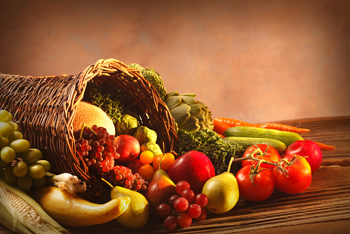 A Thanksgiving cornucopia of fruits and vegetables rests on a table in front of an old world painted background.