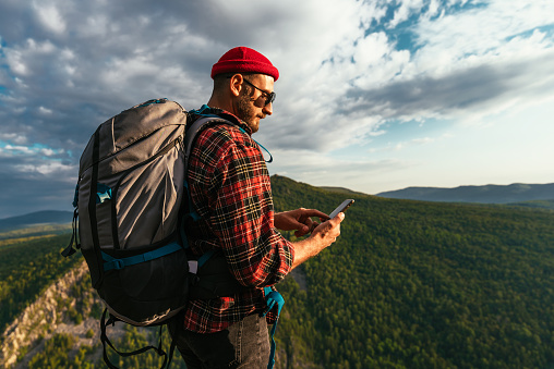 A bearded traveler with a backpack on the top of a mountain. Portrait of a traveler in a red cap and sunglasses. A tourist with a backpack stands against the background of a mountain. Copy space