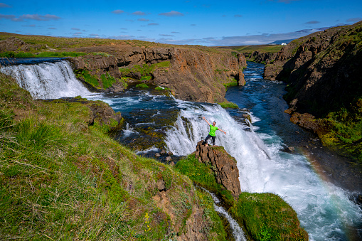 Tourist, mature woman standing on rock at the edge of Rejkjafoss waterfall on river  Huseyjarkvisl, North Iceland. She is winner, standing like statue while looking, enjoying flowing and falling water, as it is cascade, two big steps waterfall with small rainbow behind it.