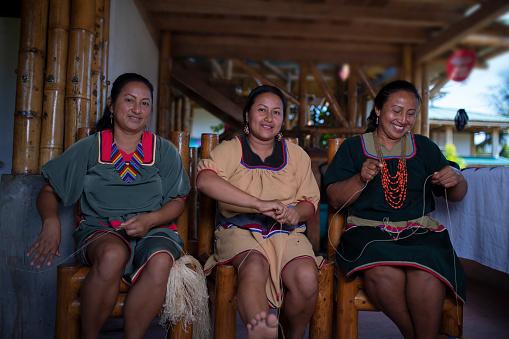 Nueva Loja, Sucumbios / Ecuador - September 2 2020: Indigenous woman of Cofan nationality with green dress smiling while weaving handicrafts sitting on a chair at her home in the Amazon rainforest