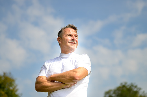 Confident senior man in white T-shirt standing with folded arms and a happy smile in a low angle view outdoors against blue cloudy sky with copyspace