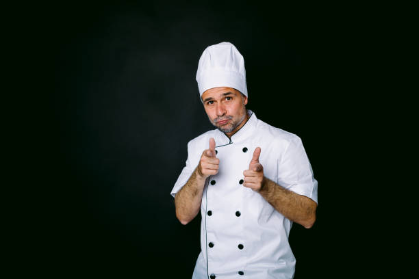 istock Cook dressed in jacket and white hat raising hands in approval, on black background. Restaurant and takeaway concept. 1344385911