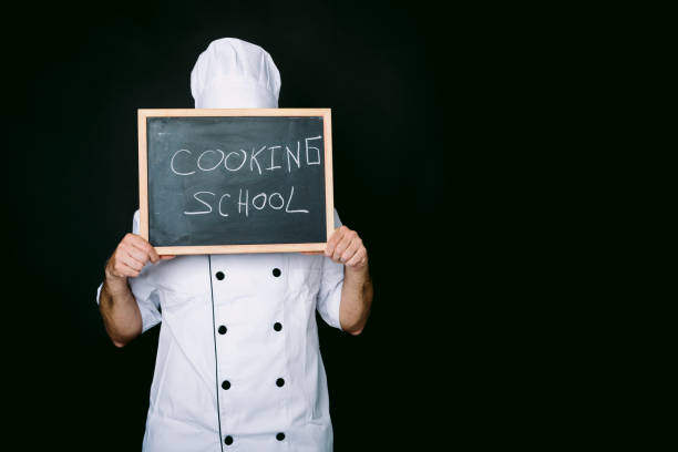istock Cook dressed in a white hat and jacket covers his face with a blackboard that reads: Cooking School on a black background. Restaurant, food and cooking school concept. 1344385949