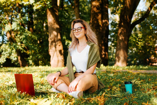 Smiling young woman freelancer meditates, relaxes during remote work outdoors. Teenage girl with glasses sits in lotus position with laptop and disposable cup of coffee on green lawn in park outdoors stock photo