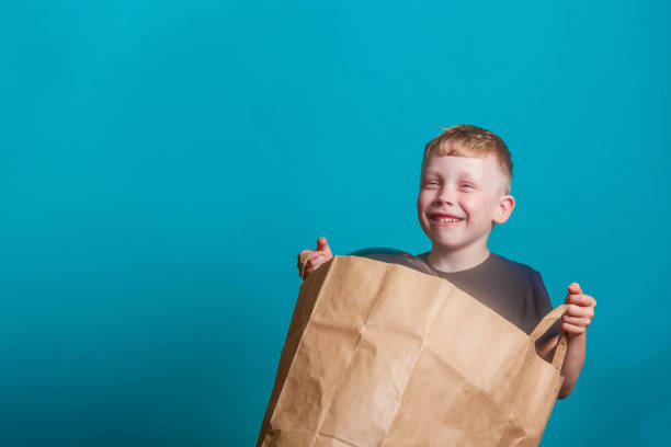 carino divertente bambino europeo che tiene in mano un sacchetto di carta con palloncini neri su sfondo blu nello studio. il bambino è sorridente e felice. spazio di copia. - child balloon selling sign foto e immagini stock