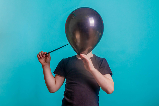 Cute funny Baby inflates and bursts a balloon on black Friday. A boy on a blue background in the Studio covered his face with a black balloon in his teeth. Copy space.