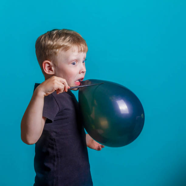 simpatico e divertente baby gonfia e fa scoppiare un palloncino il black friday. un ragazzo su sfondo blu nello studio tiene un palloncino nero tra i denti. spazio di copia. - child balloon selling sign foto e immagini stock