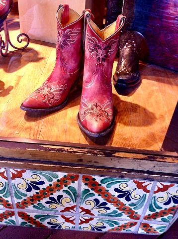 Vertical closeup photo of colourful embroidered leather boots in a store window display in Santa Fe, New Mexico. A vibrant painted tile pattern is on the lower section of the shop front.